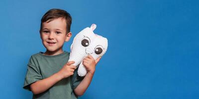 A smiling boy with healthy teeth holds a toothbrush and a plush tooth on a blue background. Pediatric dentistry. A place for your text. Panoramic photo. photo