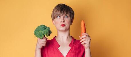 un estudio Disparo de un sonriente joven mujer con Fresco brócoli y zanahorias en su manos en un amarillo antecedentes. de cerca.. el concepto de sano comiendo. un sitio para tu texto. el panorama. foto