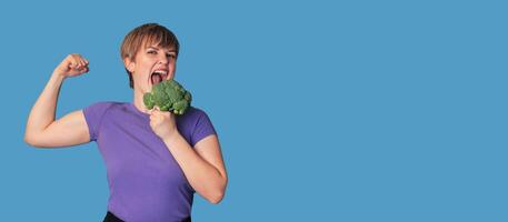 Smiling Caucasian girl with fresh broccoli in one hand and showing biceps with the other. Panoramic banner, free space. photo