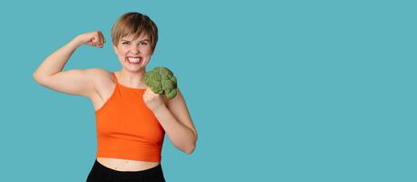 A smiling Caucasian girl with fresh broccoli in her hands as dumbbells. Panoramic banner, free space. photo