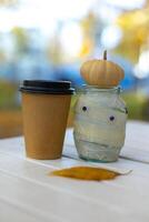 jar decor mummy with halloween pumpkin and craft glass with coffee on autumn bokeh background photo