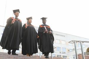 Group of happy smiling multicultural people in graduation gowns and caps with diplomas in hands graduate university outdoors in campus looking at camera. Graduation from college, university concept. photo