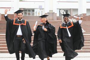 Group of happy smiling multicultural people in graduation gowns and caps with diplomas in hands graduate university outdoors in campus looking at camera. Graduation from college, university concept. photo