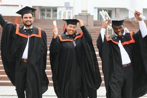 Group of happy smiling multicultural people in graduation gowns and caps with diplomas in hands graduate university outdoors in campus looking at camera. Graduation from college, university concept. photo