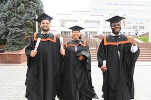 Group of happy smiling multicultural people in graduation gowns and caps with diplomas in hands graduate university outdoors in campus looking at camera. Graduation from college, university concept. photo