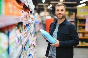 Young man in the supermarket in the household chemicals department. Large selection of products. A brunette in a glasses and a beard in a beige coat. photo