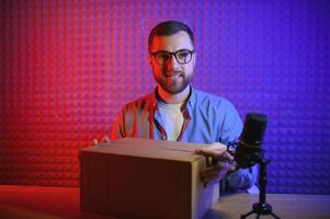 Panoramic view of young adult vlogger creating internet content, holding carton box in hands, unpacking received pack and recording media online on broadcast equipment photo