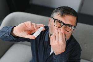 Portrait of man with a nasal spray. Sick man using nose drops while laying at the bed. Concept of treatment for allergies or the common cold, winter style. photo