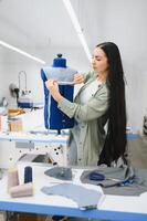 Cheerful young seamstress sits at the table with working things, in atelier photo