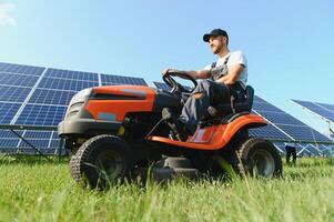 A man working at solar power station. A worker on a garden tractor mows grass on a solar panel farm. photo