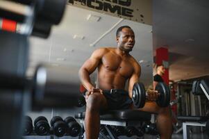 Handsome african american bodybuilder making work out with barbells at gym, copy space. Hot black guy exercising with two dumbbells, working on his muscular biceps photo