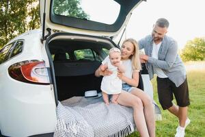 Pretty young married couple and their daughter are resting in the nature. The woman and girl are sitting on open car boot. The man is standing near them. They are smiling photo