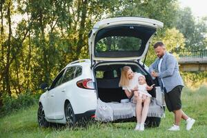 Pretty young married couple and their daughter are resting in the nature. The woman and girl are sitting on open car boot. The man is standing near them. They are smiling photo