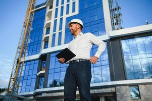 Successful engineer or architect, crane and building construction at backgrpound. Joyous businessman wearing helmet photo