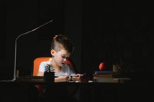 concentrated schoolboy reading book at table with books, plant, lamp, colour pencils, apple, and textbook photo