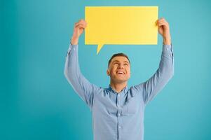 Handsome smiling man holding blank speech bubble sign isolated on light blue studio banner background with copy space photo