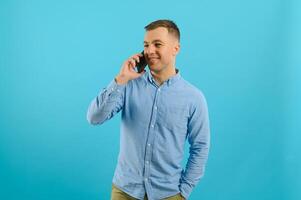 Young caucasian handsome man isolated on blue background looking at the camera and smiling while using the mobile photo
