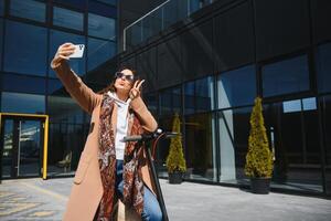A young beautiful girl is standing next to her electric scooter. She is smiling, business-trained, with modern architecture in the background. photo