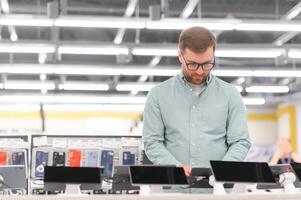 Portrait of happy satisfied young bearded man with eyeglasses testing a tablet in a tech store photo