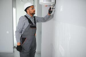 Indian plumber using screwdriver while fixing boiler or water heater, working on heating system in apartment photo