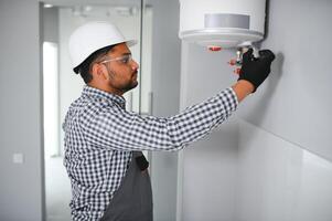 Indian plumber using screwdriver while fixing boiler or water heater, working on heating system in apartment photo
