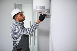 Indian plumber using screwdriver while fixing boiler or water heater, working on heating system in apartment photo