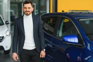 Handsome bearded buyer in casual wear in dealership, guy looks on camera while standing near car with crosed arms photo
