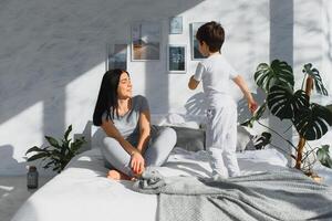 Mom playing with her child in the bedroom. Mother with son sharing good emotions while relaxing at home. Son jumping on the bed. photo