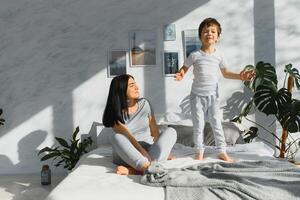 Mom playing with her child in the bedroom. Mother with son sharing good emotions while relaxing at home. Son jumping on the bed. photo