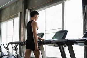 A man in a black tank top sets up a treadmill in a modern gym. His focused expression and muscular build highlight his commitment to fitness and cardio training. photo