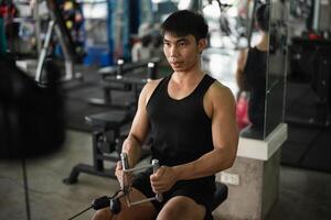 A man in a black tank top works out on a seated row machine in a modern gym. His serious expression and muscular build emphasize his dedication to fitness and strength training. photo