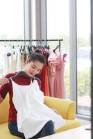 Smiling Beautiful Asian woman blogger recording on Dslr Camera for selling white dress in online shopping website on yellow sofa. Start up business and Marketing in social media concept. photo