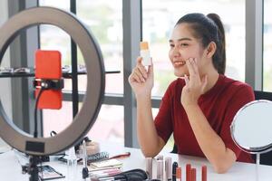 Smiling Beautiful Asian woman blogger holding Sunscreen bottle and recording on red smartphone for make up tutorial in Cosmetics website. Marketing in social media concept. photo