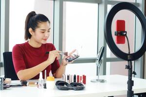 Beautiful Asian woman blogger hand holding Eye shadow box and recording on red smartphone for make up tutorial in Cosmetics website. Marketing in social media concept. photo