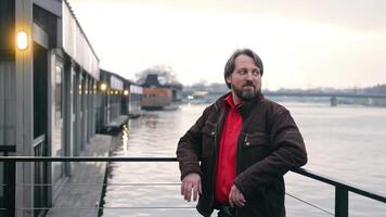 A man with a beard stands by the riverside, leaning on a railing, with modern buildings and the river in the background at dusk. video