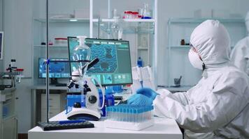 Chemist in protection suit examining various bacteria holding petri dish in laboratory typing on pc. Doctors analysing vaccine evolution using high tech researching diagnosis against covid19 virus. video