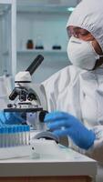 Vertical . Close up of scientist looking through a microscope wearing ppe suit in equipped lab. Team examining virus evolution using high tech for scientific research of vaccine development against covid19. video