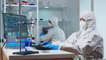 Team of biologists in coverall working with microscope and typing on computer sitting in laboratory. Scientists examining vaccine evolution using high tech researching diagnosis against covid19 virus video