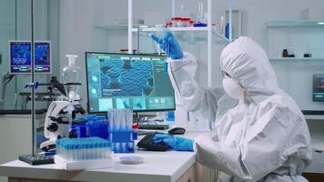 Biochemists in coverall typing at pc evolution of vaccine composition in equipped laboratory analysing test tube with sample. Doctors examining virus evolution using high tech researching diagnosis video