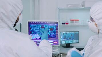 Scientists coworkers in coverall discussing about virus evolution in equipped laboratory pointing on computer desktop. Doctors examining vaccine evolution using high tech researching diagnosis video