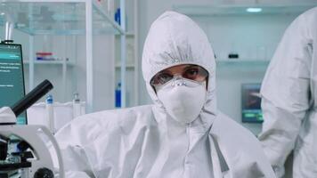 porträtt av trött forskare i overall ser på kamera Sammanträde i modern utrustad laboratorium. team av doktorer granskning virus Evolution använder sig av hög tech och kemi verktyg för vaccin utveckling. video