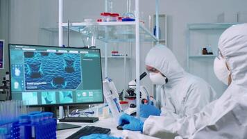 Chemist doctor in ppe suit working at pc while lab technician using microscope. Team of scientists examining vaccine evolution with high tech for researching treatment against covid19 virus video