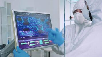 Chemist in ppe suit explainig to doctor vaccine development in modern equipped laboratory pointing on computer desktop. Team of doctors examining virus evolution using high tech researching diagnosis video