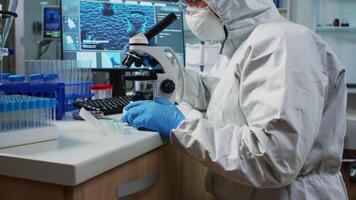 Scientist in protection suit looking through a microscope in equipped lab. Team of chemists examining virus evolution using high tech for scientific research of vaccine development against covid19. video