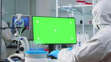 Scientist man wearing protection suit typing on computer with green mockup in modern equipped lab. Team of microbiologists doing vaccine research writing on device with chroma key, isolated display. video