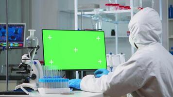 Doctor with coverall working at computer with green screen in modern equipped lab. Team of microbiologists doing vaccine research writing on device with chroma key, isolated, mockup display. video
