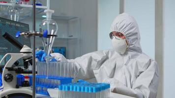 Technician in ppe suit doing experiment checking test tube in equipped lab typing results on pc. Scientist in coverall working with various bacteria tissue and blood samples for antibiotics innovation video