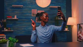 African employee taking a selfie using mobile phone smiling at camera sitting at desk in living room late at night. Black woman relaxing using modern technology network wireless doing overtime for job video