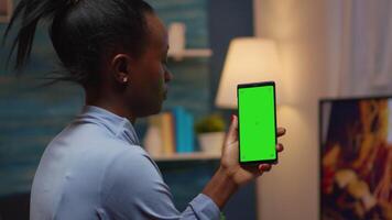 Housewife holding smartphone with chroma screen on hand looking at mockup. Reading on green screen template chroma key isolated mobile phone display using techology internet sitting on cozy couch video