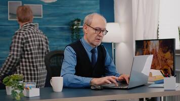 Focused old entrepreneur checking graphics writing on laptop working from home drinking coffee. Retired employee using modern technology reading typing, searching while senior wife sitting on couch video